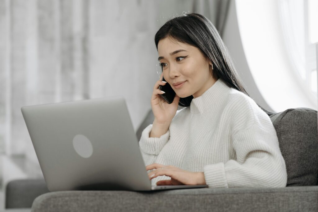 Woman multitasking from home using a laptop and smartphone on a sofa. Viewing website designed by JHC Web Design.